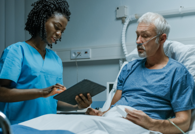Eine Pflegefachfrau mit einem Tablet-Computer spricht mit einem älteren Patient im Krankenhaus.