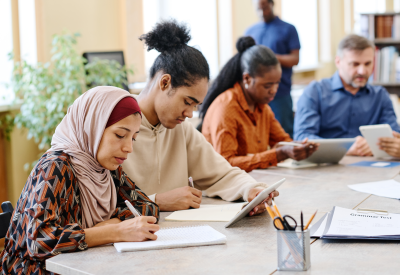 Gruppe ethnisch gemischter Menschen, die an einem Tisch sitzend mit Hilfe digitaler Tablets Sprachunterricht für Einwanderer erteilt