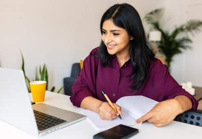 Junge erwachsene indische Studentin, die sich Notizen macht, während sie einen Laptop zu Hause benutzt. Ethnische Millennial-Frau, die online lernt und einem virtuellen Videoanruf zuhört. Business und Bildung Konzept.