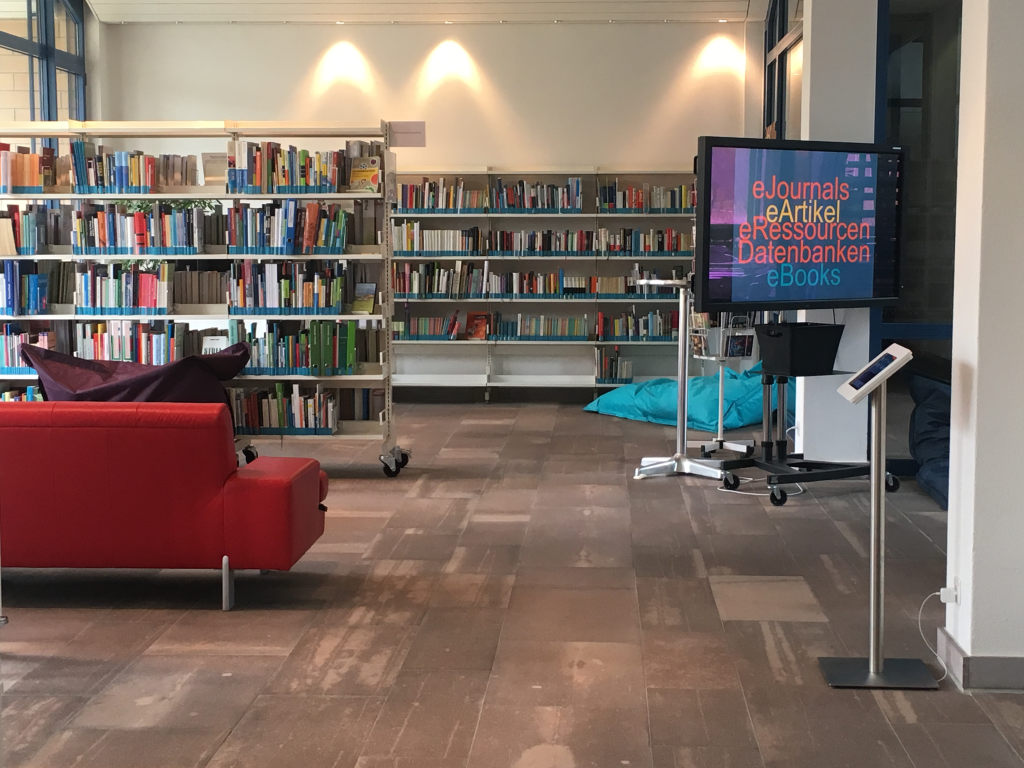 Ansicht eines Teilbereiches der Bibliothek: Bücher, Screen mit Zugang zu den elektroischen Medien, ein iPad für die Recherche, ein rotes Ledersofa zum verweilen.