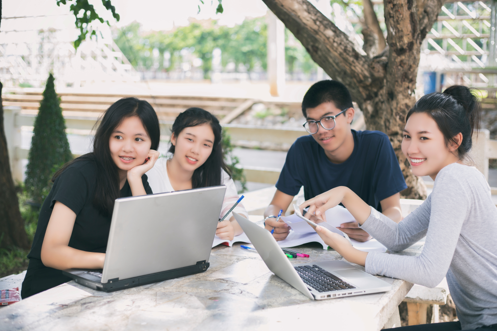 Asiatische Studentengruppe mit Tablet und Notebook, die Ideen für die Arbeit auf dem Rasen des Campus austauschen